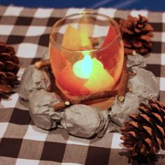 a lit candle sitting on top of a checkered table cloth with pine cones around it