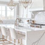 a white kitchen with marble counter tops and gold accents on the cabinets, along with chandeliers