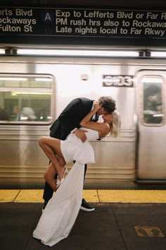 a man and woman kissing in front of a train at a subway station, with their arms around each other