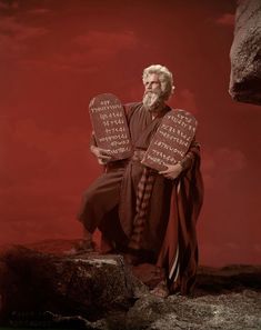 an old man holding two wooden signs on top of a rocky cliff with red sky in the background