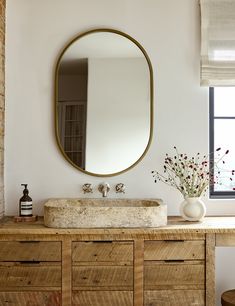 an oval mirror hangs above a sink in a bathroom with wooden cabinets and drawers, along with a vase filled with flowers