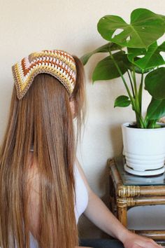 a woman with long hair wearing a crocheted headband sitting in front of a potted plant