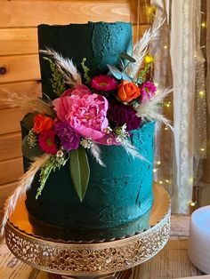 a blue cake with flowers and feathers on top sits on a gold platter next to a candle