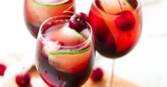 two glasses filled with liquid and cherries on top of a cutting board next to each other