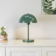 a green table lamp sitting on top of a white dresser next to a potted plant