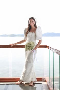 a woman in a wedding dress standing on a balcony next to the water holding a bouquet