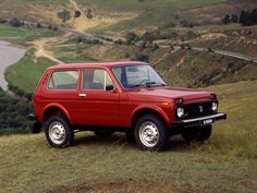 an old red car parked on top of a grass covered hill next to a river