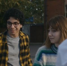 two young people standing next to each other in front of a brick building and trees