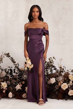 a woman standing in front of flowers wearing an off the shoulder purple dress with thigh high slit