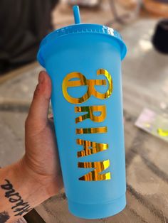 a person holding up a blue cup with gold lettering on the side and a straw in it