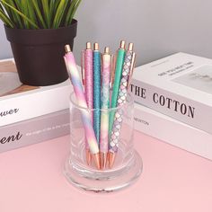 there are many different colored pens in the glass cup on the table next to books