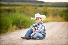 a young boy sitting on the side of a dirt road wearing a cowboy hat and jeans