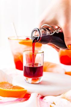 someone pouring red liquid into a glass with an orange slice on the table next to it