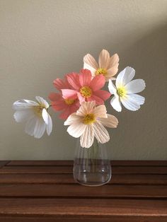 three flowers in a glass vase on a wooden table with a white wall behind them