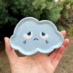 a hand holding a blue cloud shaped dish with eyes on it's face, in front of some bushes