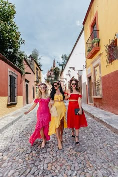 three women in colorful dresses walking down a cobblestone street with their arms around each other