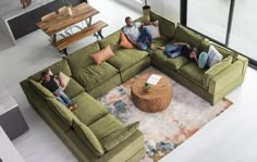 three people sitting on a green couch in a living room with a coffee table and large windows