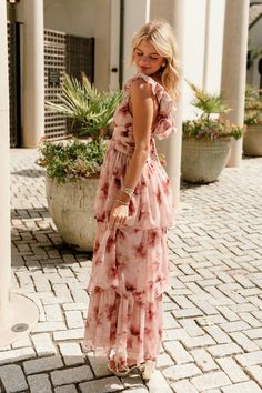 a woman in a pink dress standing on a brick walkway with potted plants behind her