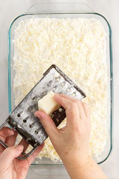 someone using a grater to scoop cheese into a casserole dish with one hand