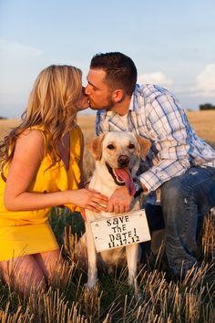 a man and woman sitting in the grass with a dog next to them, kissing