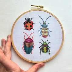 a hand is holding up a cross stitch hoop with three bugs on it and one beetle in the middle