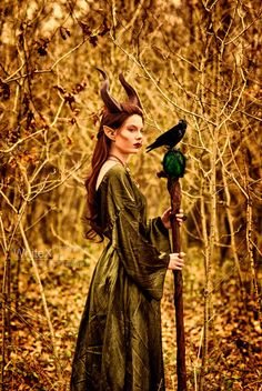 a woman with horns holding a stick and a bird on it's head in the woods