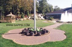 a flag pole in the middle of a garden with flowers around it and a water fountain behind it