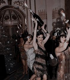 two women in flappers hold up champagne bottles as they dance with other people behind them