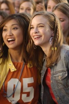 two girls are smiling and standing in front of a group of other women, one is wearing an orange shirt