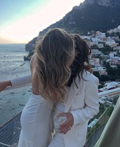 two women standing next to each other on top of a building near the ocean and mountains