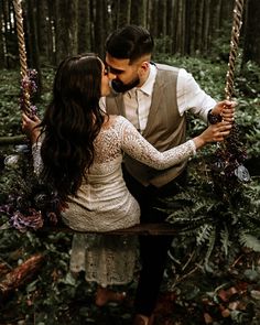 a man and woman sitting on a swing in the woods kissing each other while holding hands