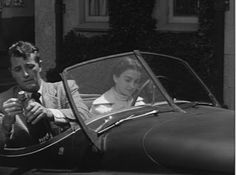 a man and woman sitting in an old fashioned car, one is holding the steering wheel