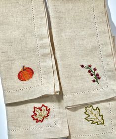 three napkins with embroidered autumn leaves and pumpkins on them are sitting on a white surface