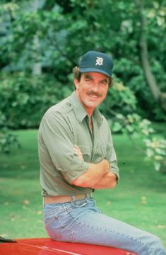 a man sitting on top of a red car wearing a baseball cap and blue jeans