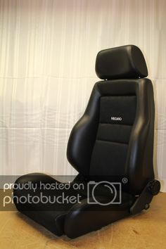 a black leather car seat sitting on top of a wooden floor next to a white curtain