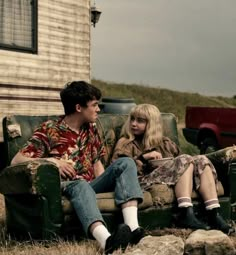 a young man and woman sitting on a couch in front of an old house with a red pickup truck behind them