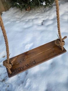 a wooden sign hanging from a rope on top of snow covered ground next to a christmas tree