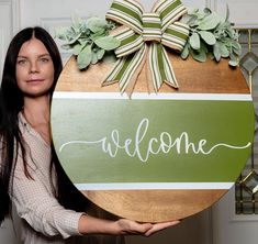 a woman holding up a welcome sign in front of a door with greenery on it