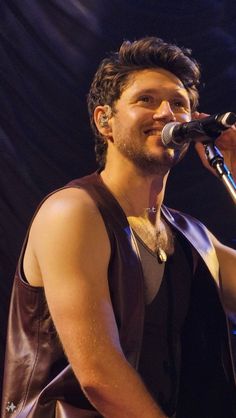 a man singing into a microphone while wearing a leather vest and holding a guitar in his right hand