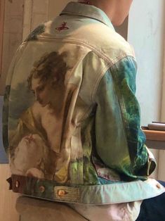 a person wearing a jean jacket with a painting on it's back, sitting in front of a window