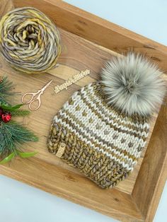 a knitted hat with two pom - poms and scissors on a wooden tray