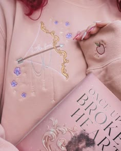 a woman is holding a book in her hands and wearing a pink sweater with an embroidered design on it