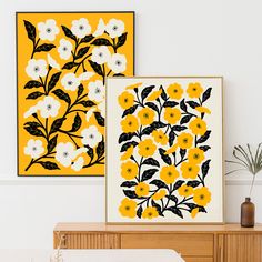 two yellow and black floral paintings on a white wall next to a wooden table with a plant