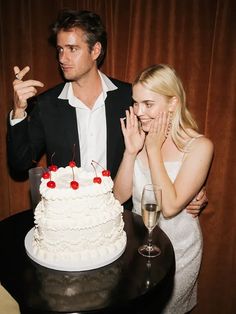 a man standing next to a woman in front of a white cake with cherries on it