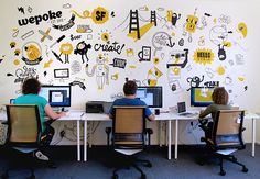 two people sitting at desks in front of computer monitors with yellow and black doodles on the wall behind them