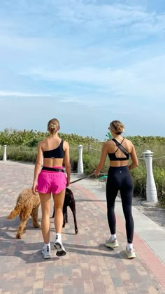 two women walking their dogs on a brick path near the ocean and grass, with one woman in black sports bra top and pink leggings