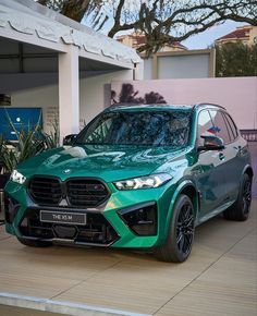 a green suv is parked in front of a pink wall and some plants on the sidewalk