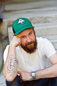 a man with a beard wearing a green hat and sitting on some steps, looking at the camera