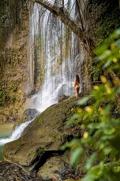 a woman standing on top of a rock next to a waterfall in the woods with water coming out of it