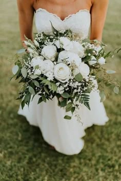 a bride holding a bouquet of white flowers and greenery on her wedding day in the grass